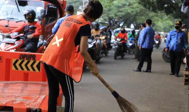 Seorang sedang menyapu jalan raya di Jakarta sambil memakai rompi jingga bertuliskan pelanggar PSBB